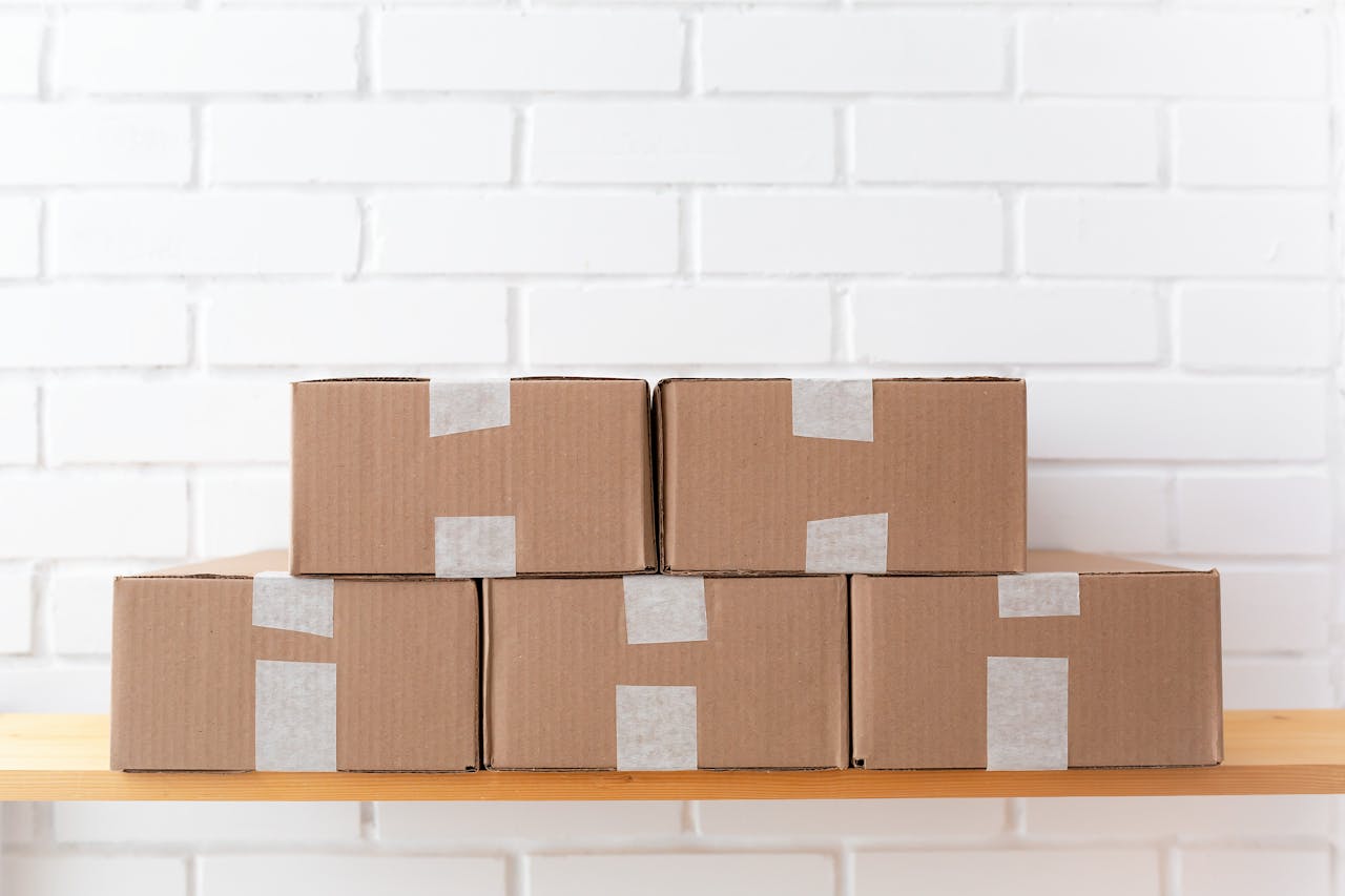 gallery-2 A neat stack of cardboard boxes taped and placed on a wooden shelf against a white brick wall.
