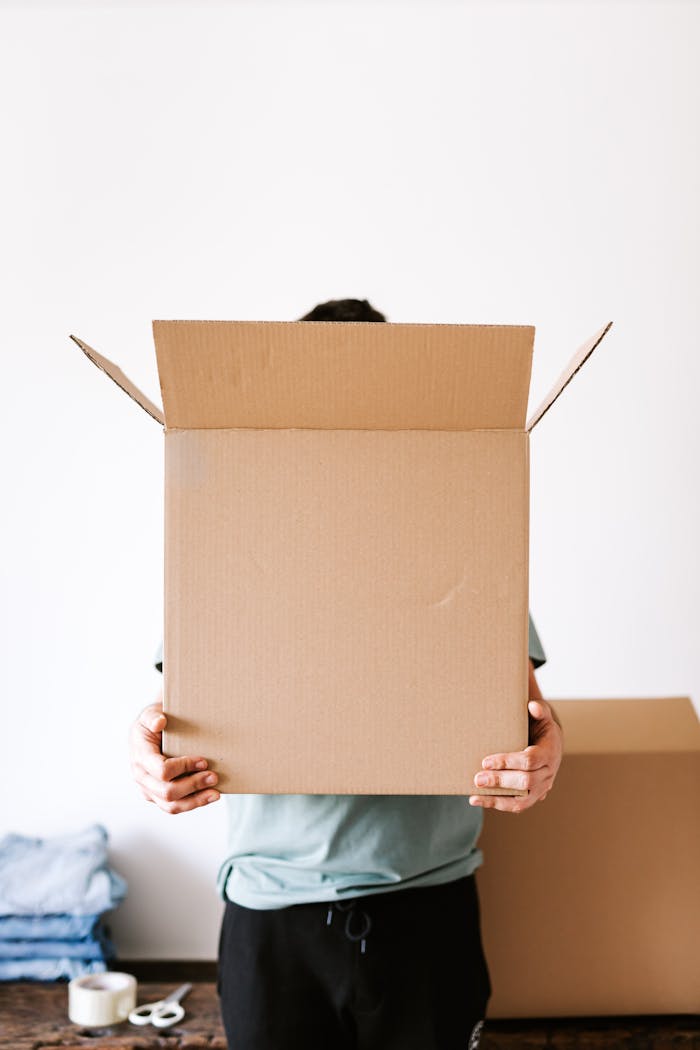 gallery-5 Anonymous man carrying opened carton box before packing belongings and relocation while standing against white wall and stacked clothes